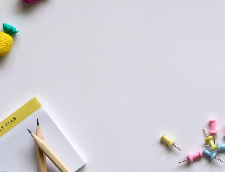 Flat lay of desk items including a planner, pencils, erasers, and push pins on a white background.