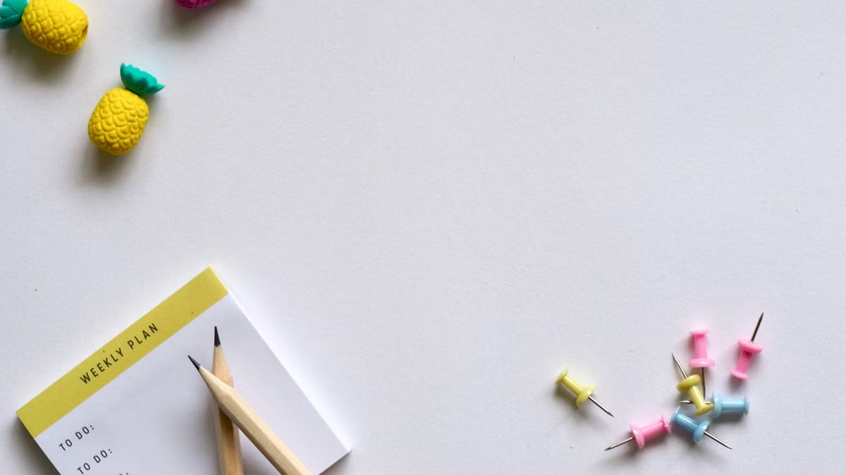 Flat lay of desk items including a planner, pencils, erasers, and push pins on a white background.